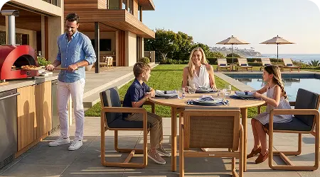 Family dining at a teak outdoor table beside a pool while a man prepares pizza in a red outdoor oven, with ocean views beyond the patio.