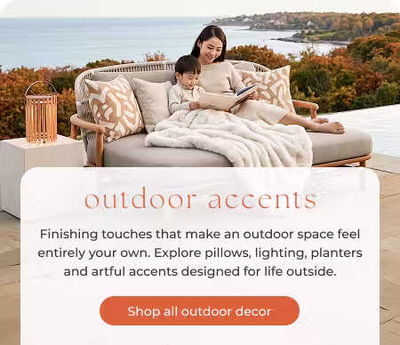 Mother and child reading on an outdoor sofa with a plush blanket, throw pillows and lantern overlooking a fall coastline.