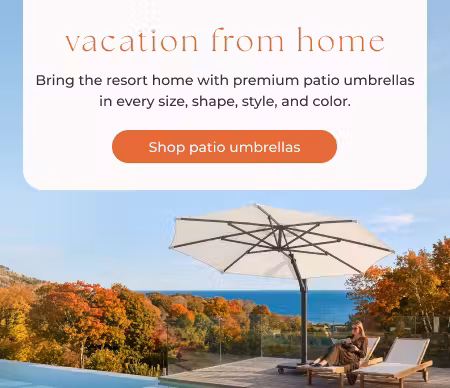 Woman reading beneath a large cantilever patio umbrella beside chaise lounges on an autumn coastal terrace.