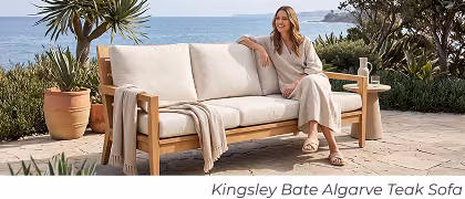 Woman sits on cushioned teak sofa. Image Text: Kingsley Bate Algarve Teak Sofa